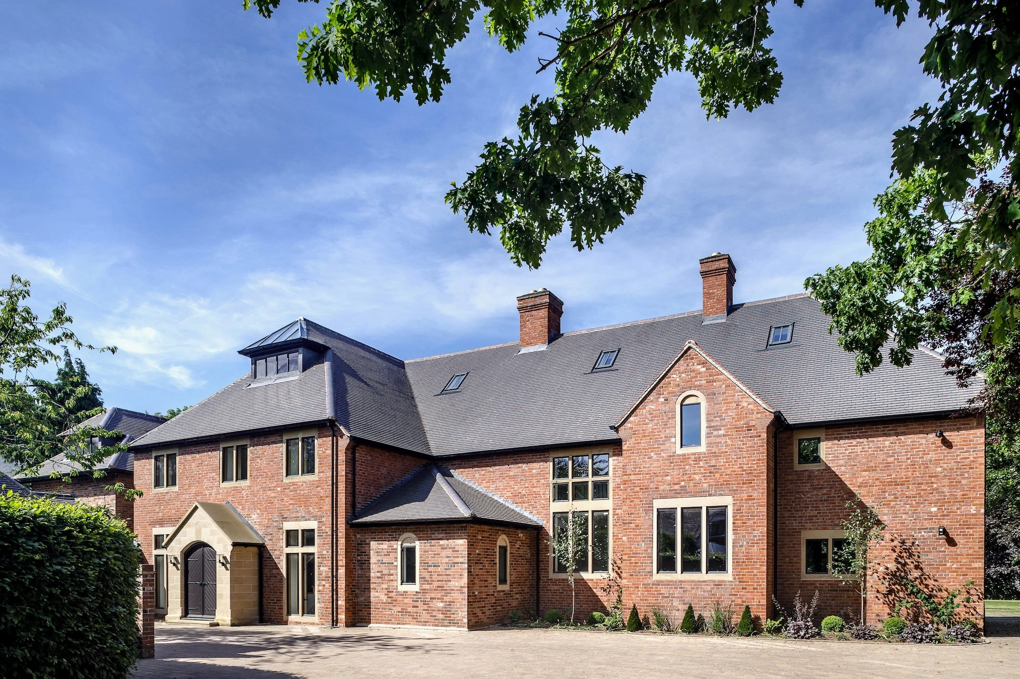 Exterior front of large house with brick walls and slate roof with paved driveway in front and trees surrounding