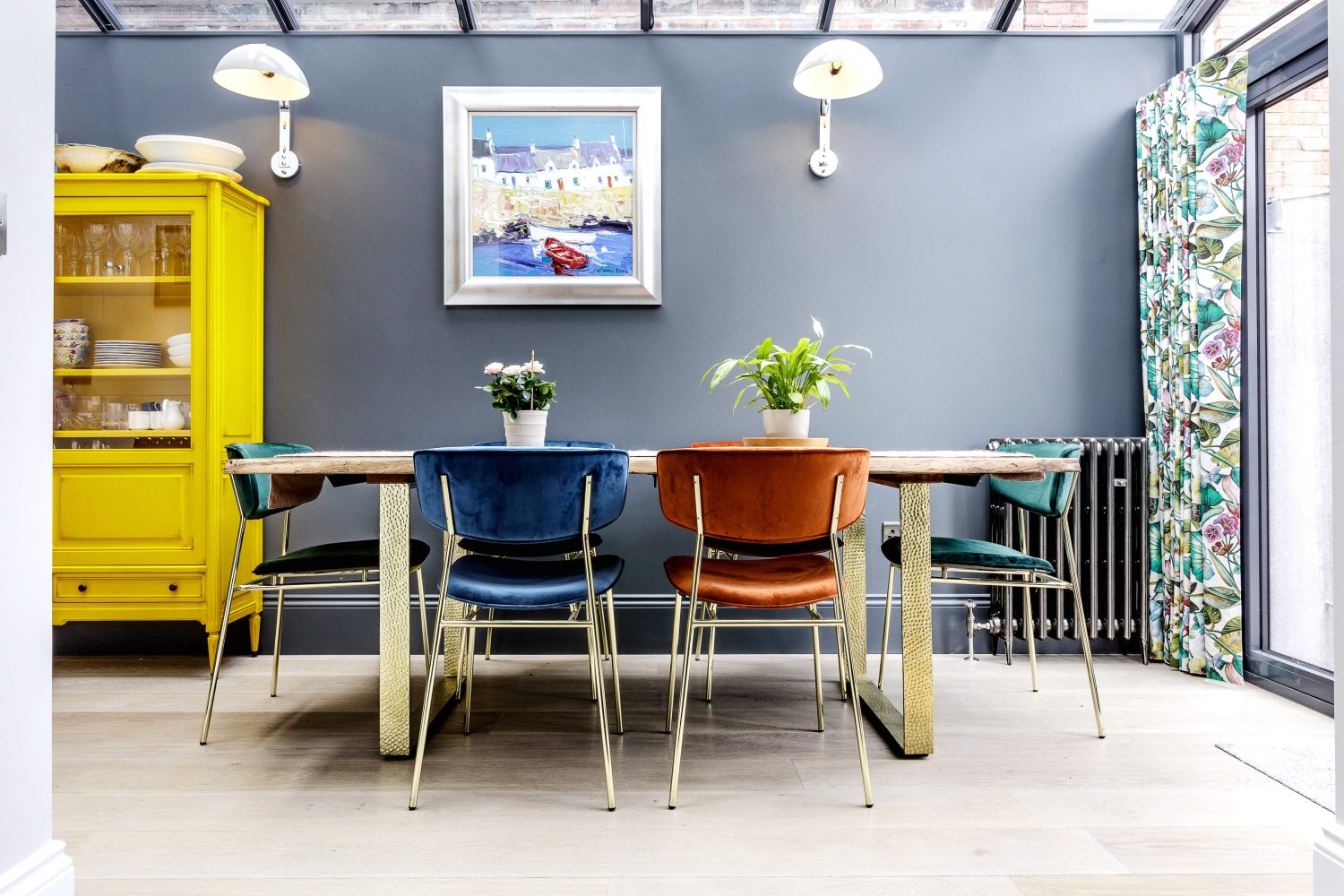 Dining table with jewel coloured chairs surrounded by a yellow cabinet and framed artwork