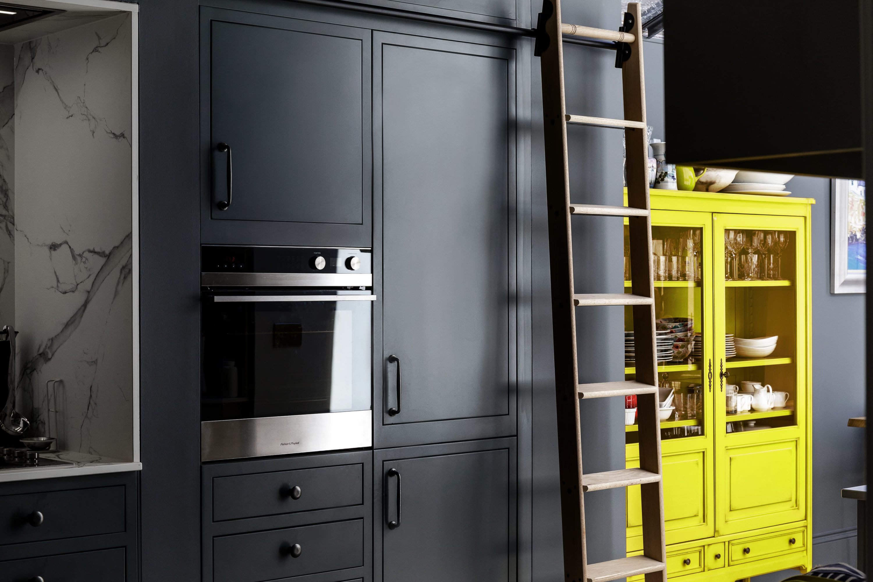 Wooden sliding ladder in dark blue kitchen with yellow cabinet beside