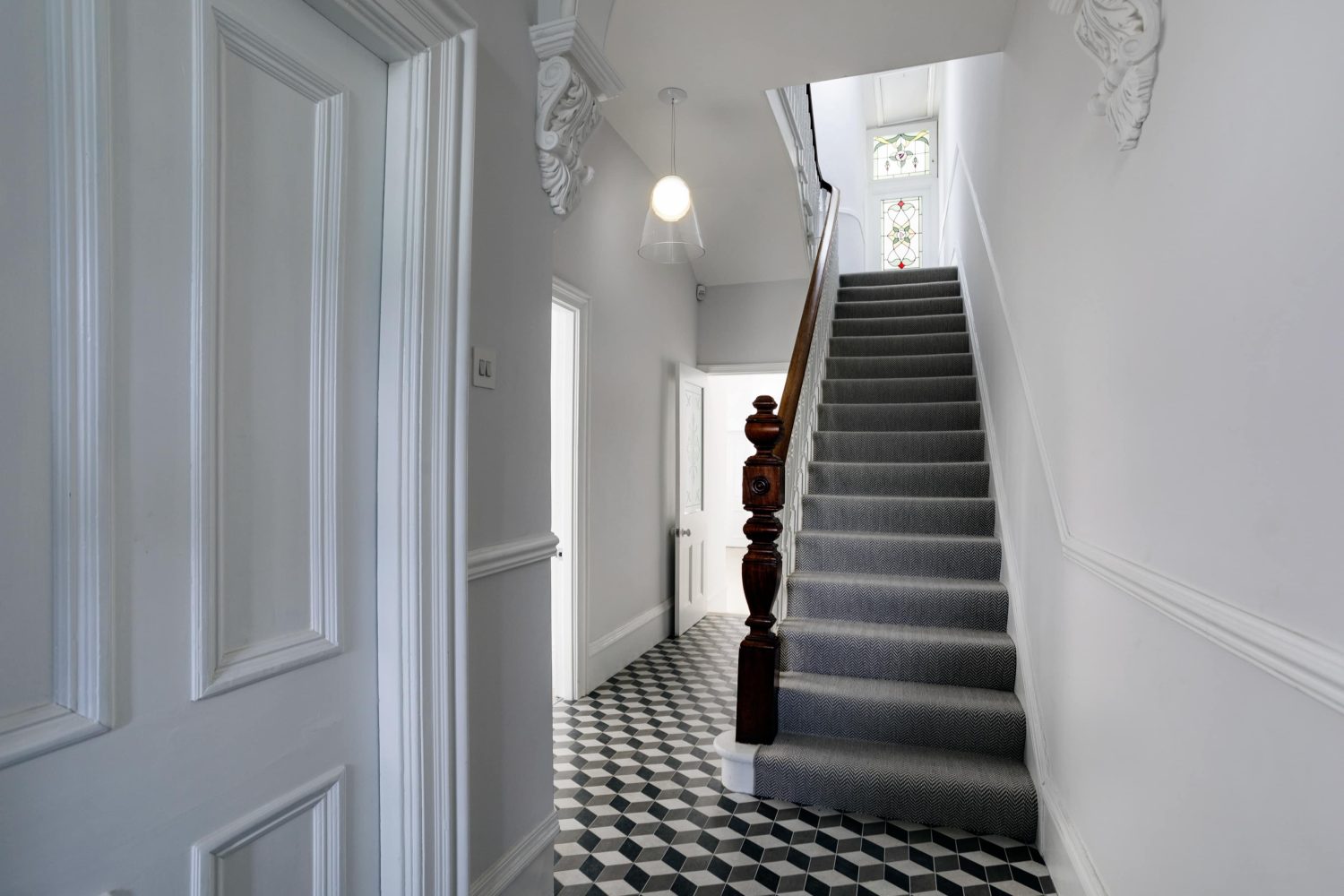 Hallway with white walls and white and grey geometric flooring. Grey carpeted staircase with a dark wooden banister