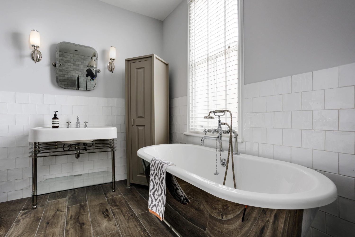 Bathroom with white and silver bath and sink. A tall beige cupboard in the corner with white tiled walls surrounding