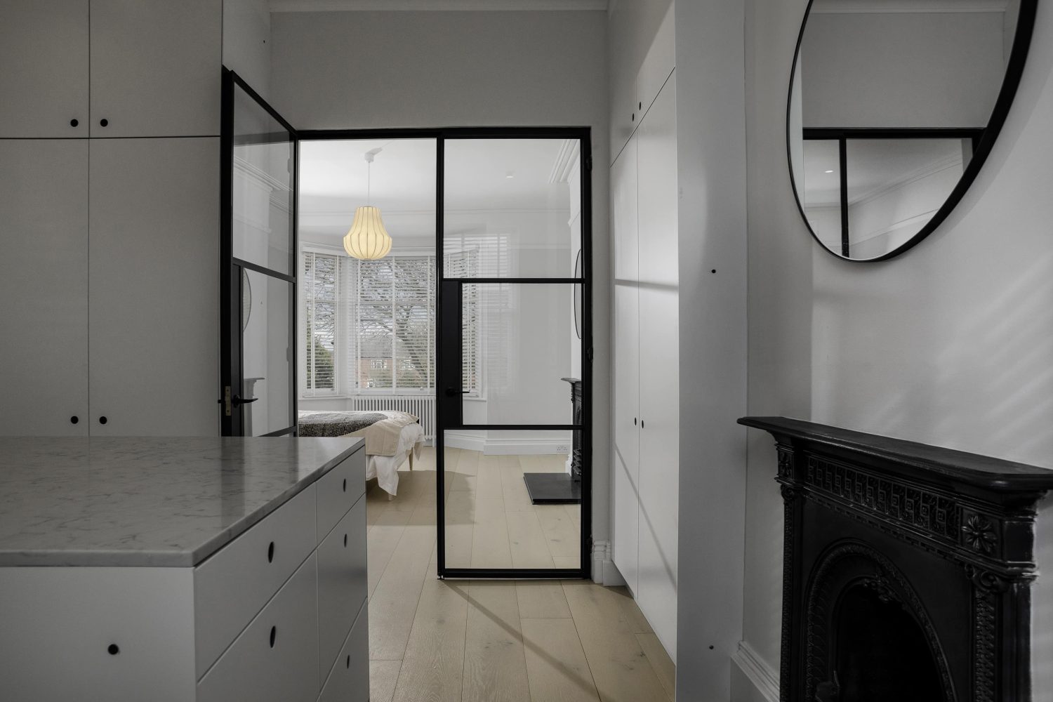 Dressing room with white built-in wardrobes and drawer unit in the centre across from black fireplace and circular mirror with Crittall-style doors behind
