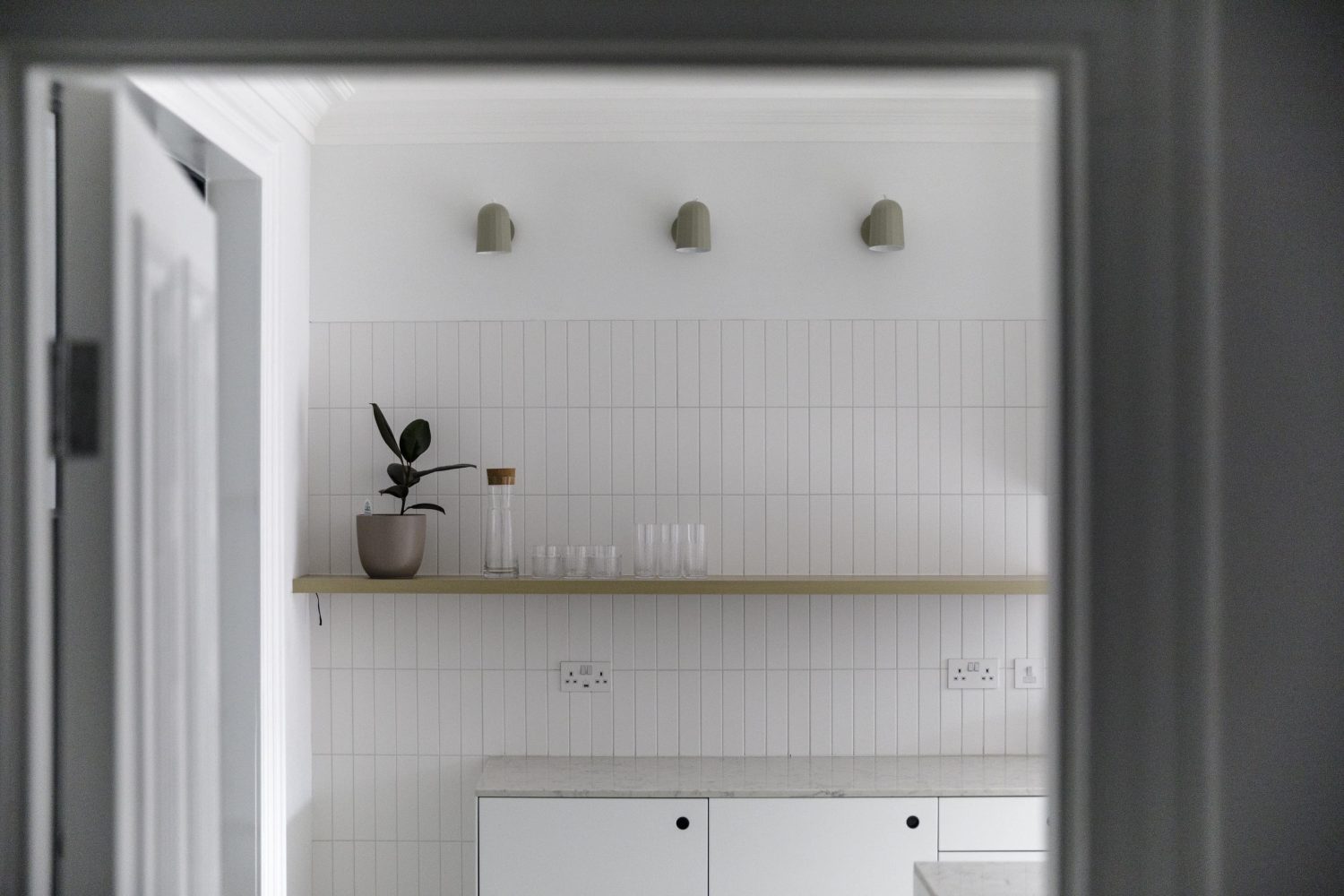 View through doorway to white tiled wall with a long shelf with glassware on top and three wall lamps above