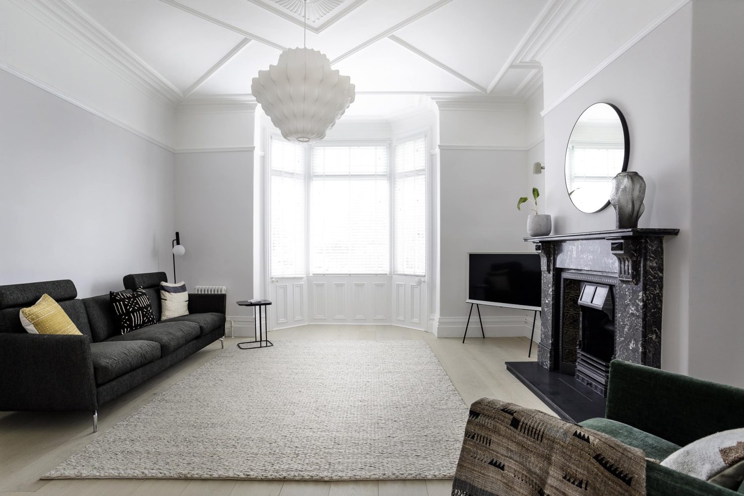 Living room with white ceiling and light grey walls and bay window. Dark grey sofa opposite black marble fireplace and a large pendant light hanging above cream rug on the floor