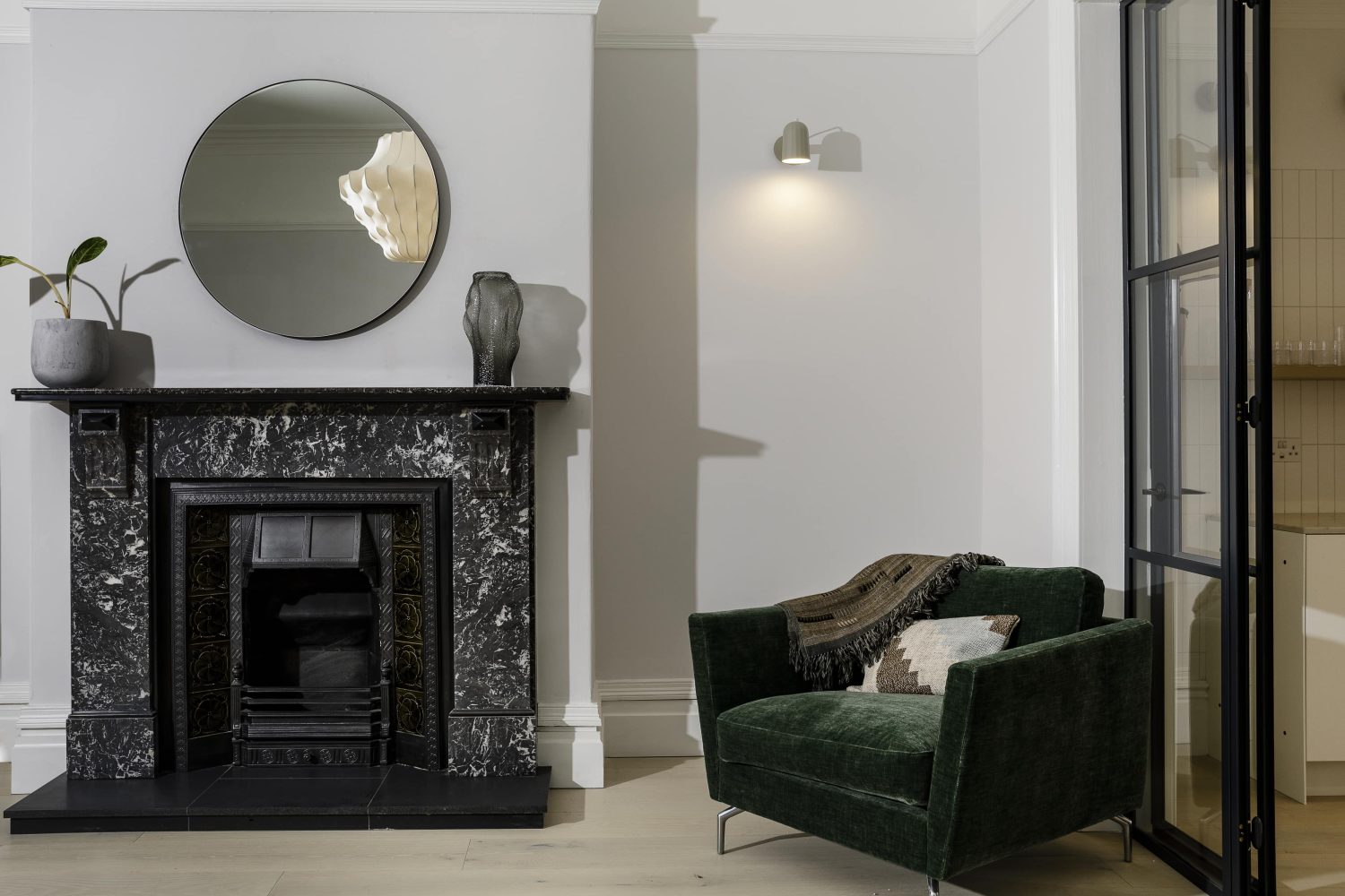 Black fireplace with a black marble mantlepiece and a circular mirror on the wall above. Dark green armchair to the right beside Crittall-style door
