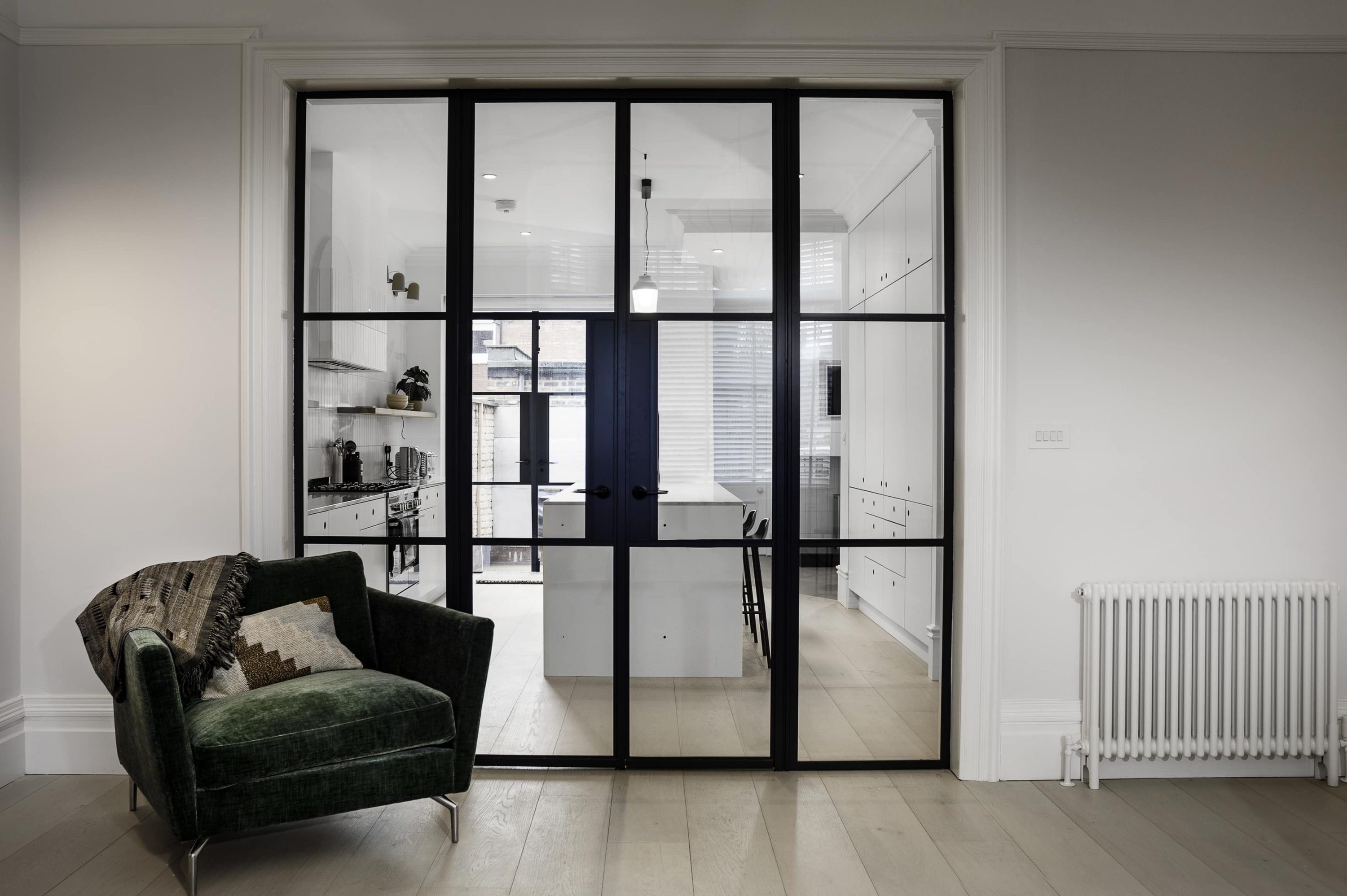 Crittall-style doors with a dark green armchair beside them looking through to a white kitchen