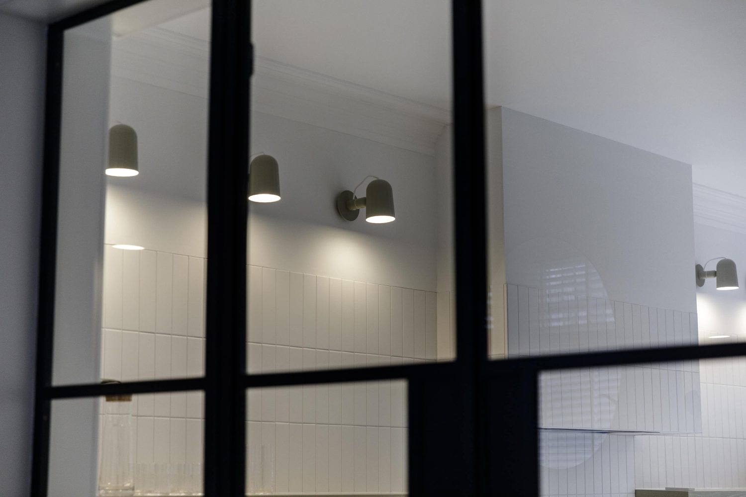 View through a Crittall-style glazed door to a white tiled wall with wall lamps lining it