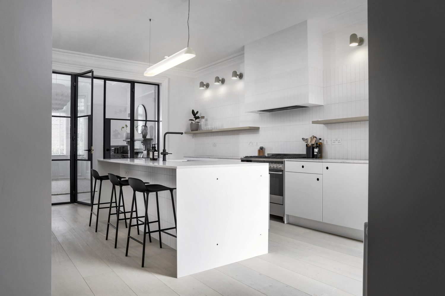 Kitchen with white tiled walls and island in the centre with black barstools and Crittall-style doors to the left
