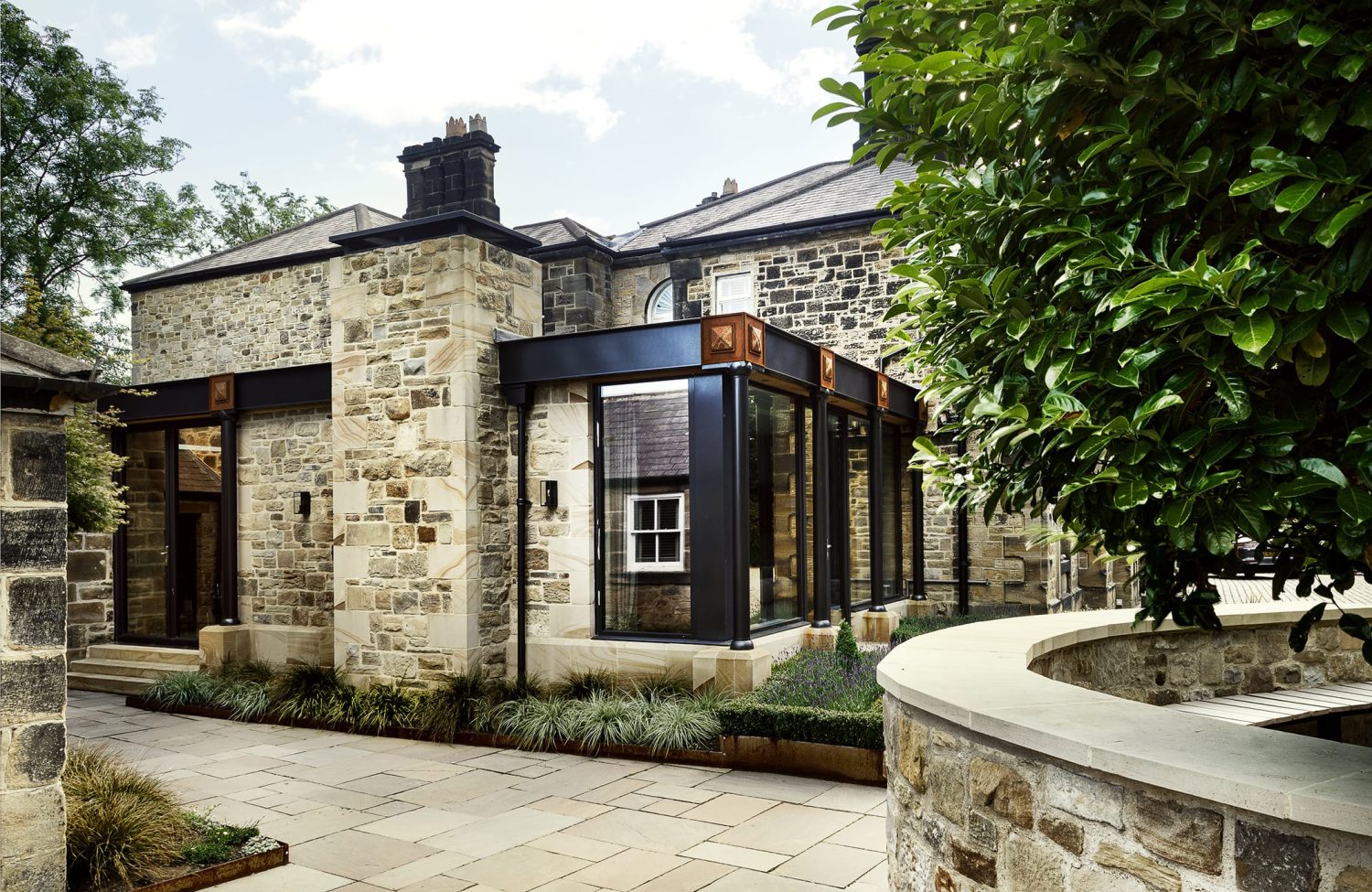 Exterior side of stone built house with black framed windows featuring copper accents and flower beds surrounding