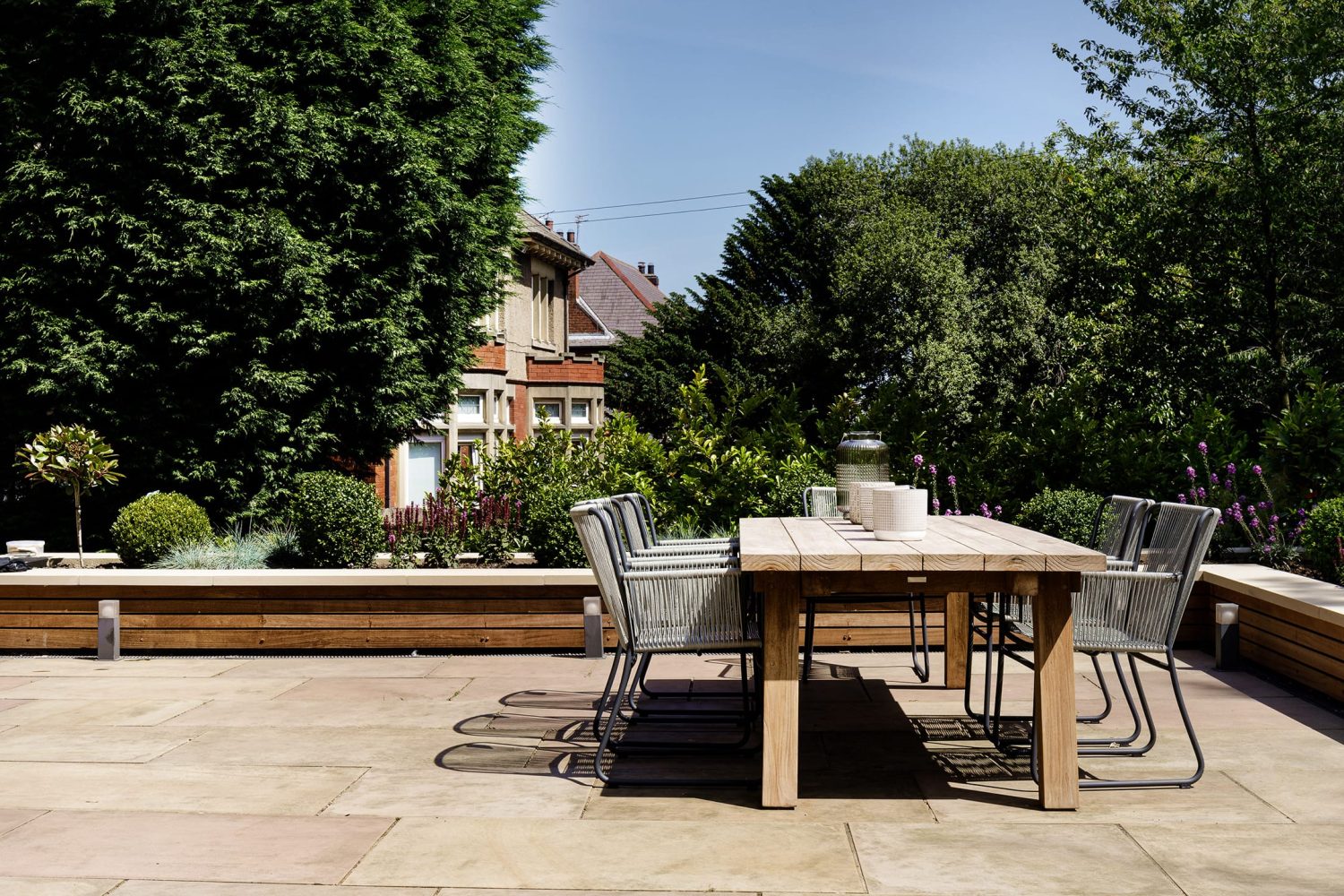 Outdoor seating area with wooden table and grey chairs on stone paved floor with trees behind