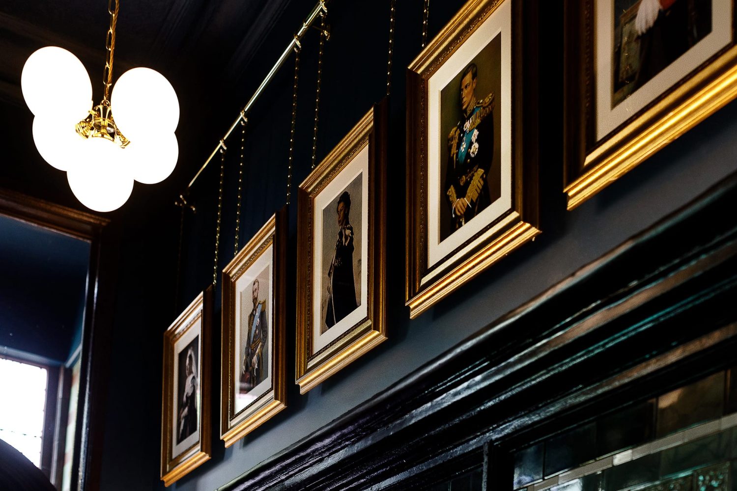 Gold framed portraits hanging from chains on a dark blue wall with pendant light hanging to the left