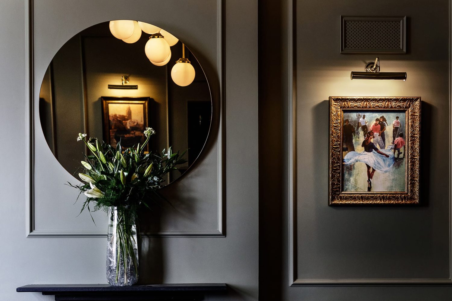 Vase of flowers in front of circular mirror on a muted blue wall with a gold framed painting and wall lamp to the right