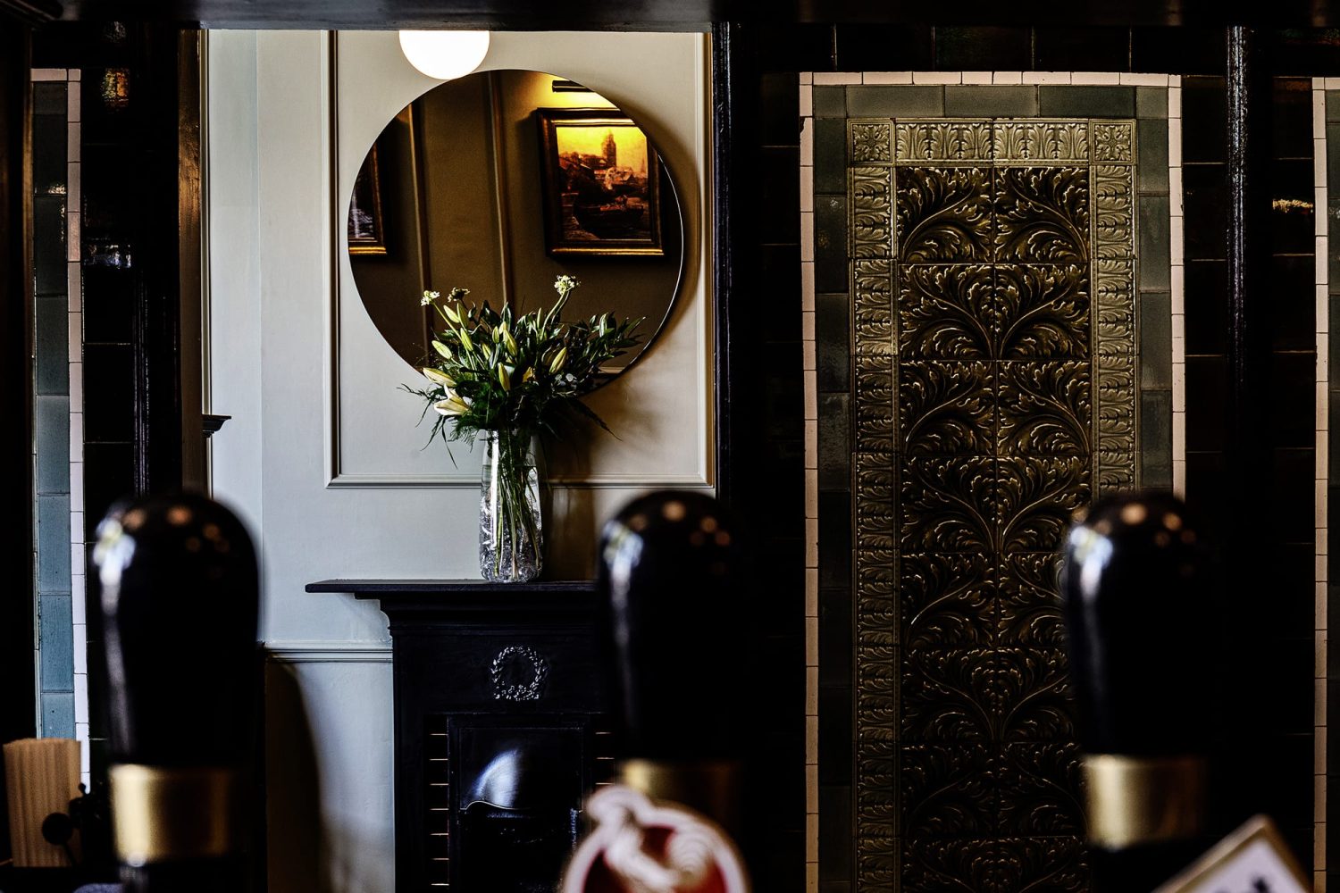 Doorway with ornate green tiles to the right looking through to small black fireplace with circular mirror on wall above