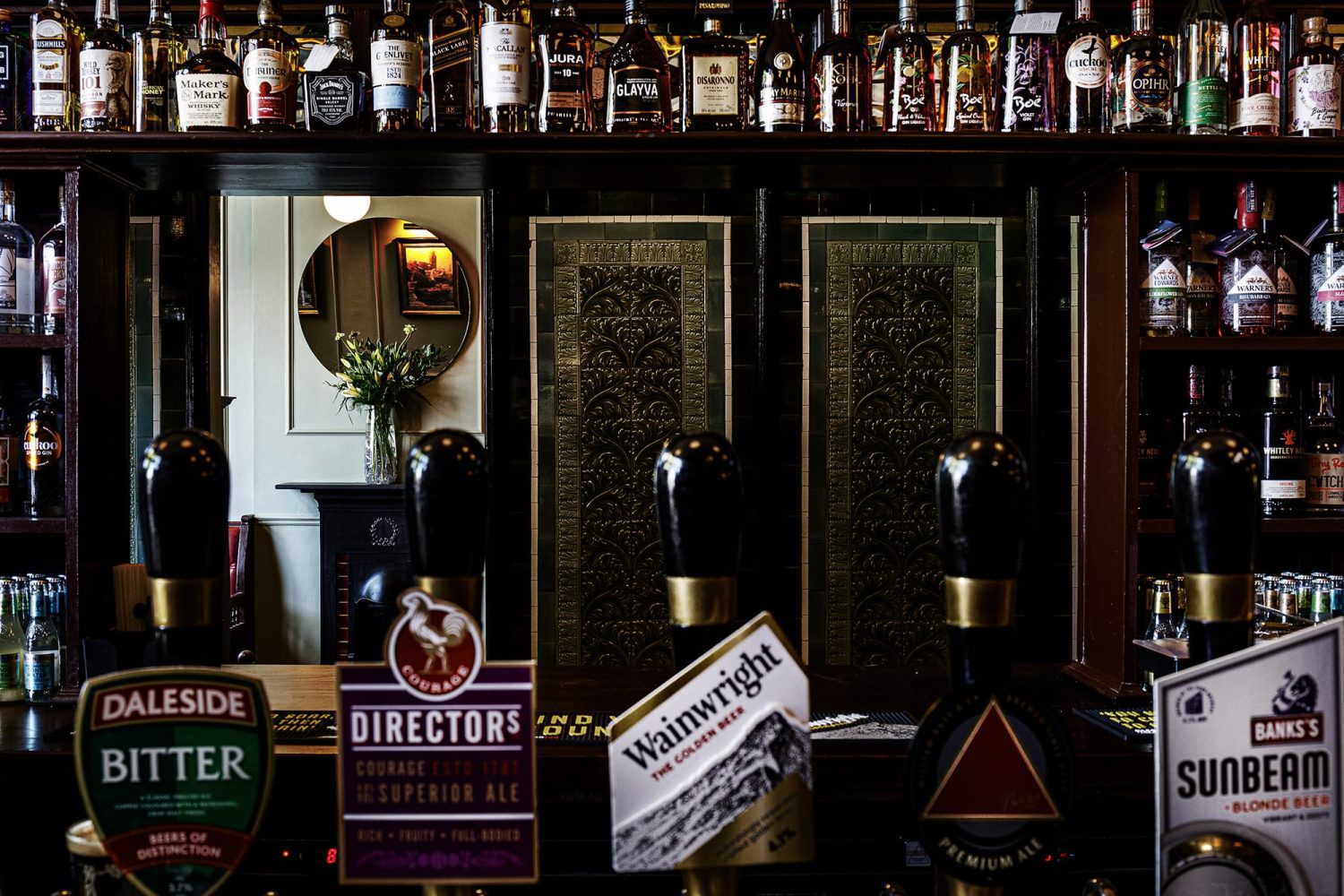 Bar with shelving holding bottles and window looking through to green tiled wall