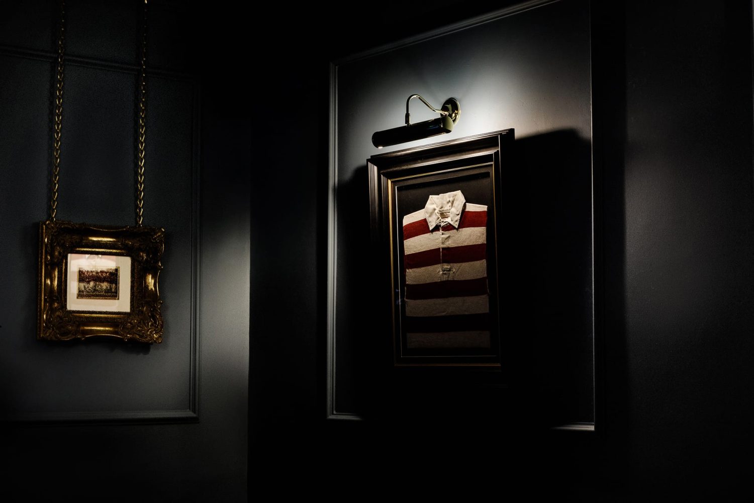 Red and white striped shirt framed on a wall with a wall lamp glowing above next to a gold framed image hanging from gold chains