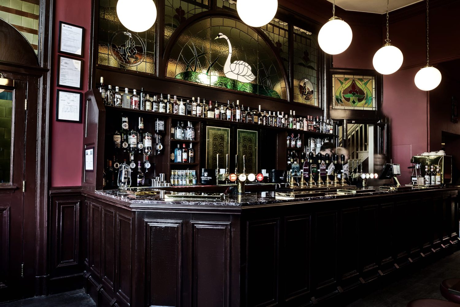 Dark brown bar with shelving behind holding bottles and a large stained glass window featuring a swan above