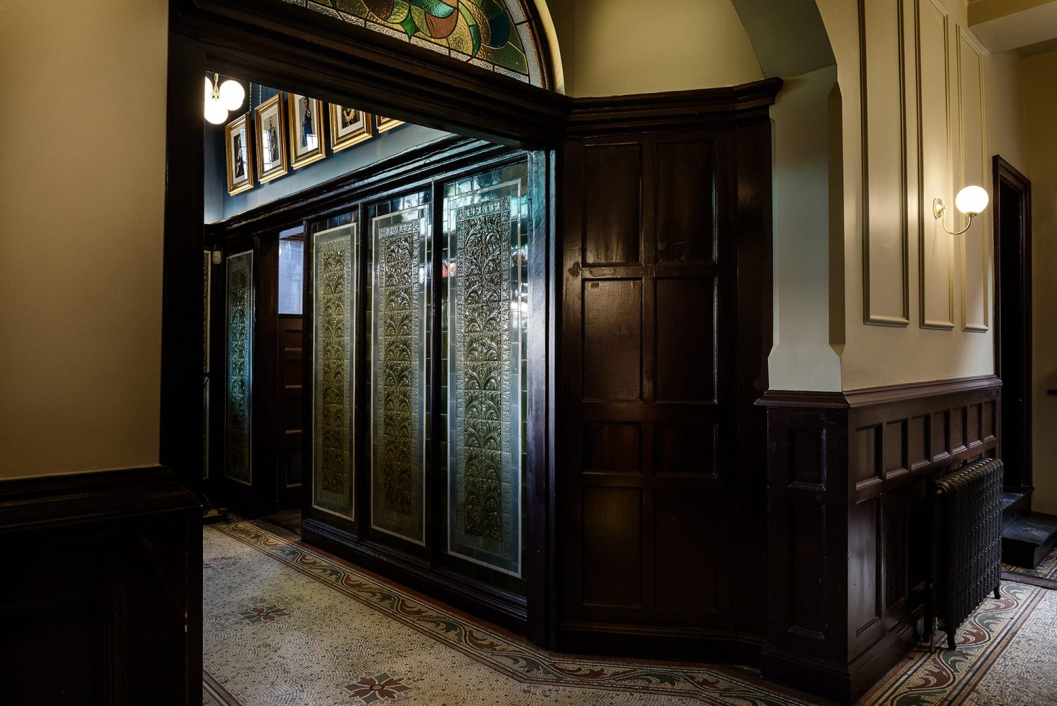 Hallway with cream walls and dark brown wooden panelling. Wall to the left featuring green tiles with ornate design