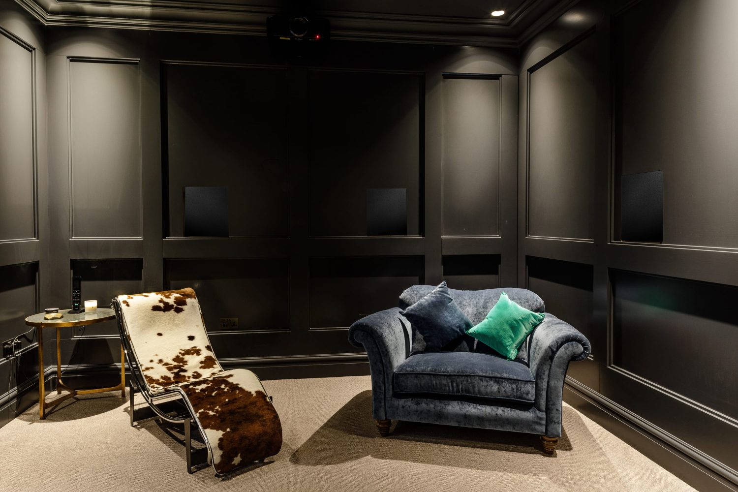 Room with dark grey panelled walls with a projector attached at the top. A blue armchair next to a cow print lounge chair