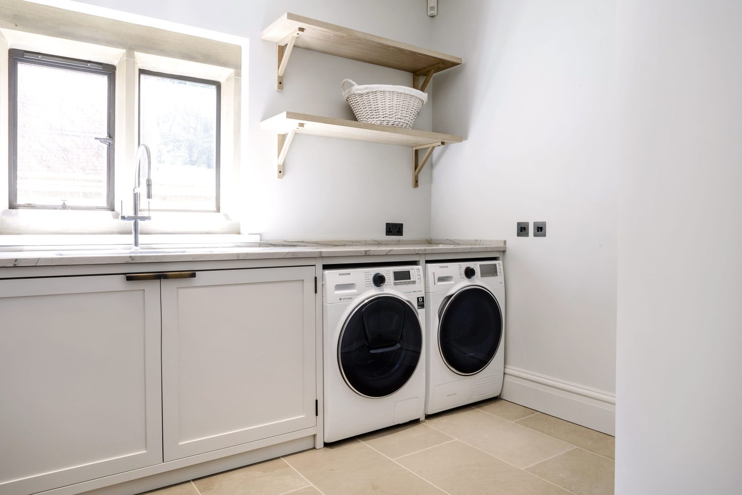 Utility room with light grey cabinets and sink next to washing machines with wooden shelves on wall above