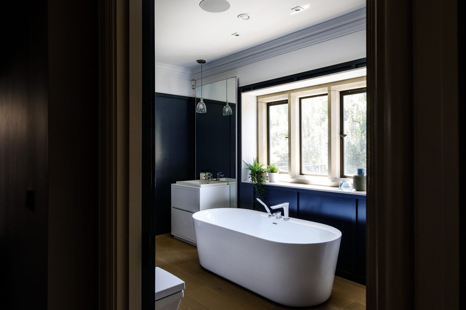 Bathroom with dark blue walls and white bath in the centre with white sink unit and tall mirror to the left