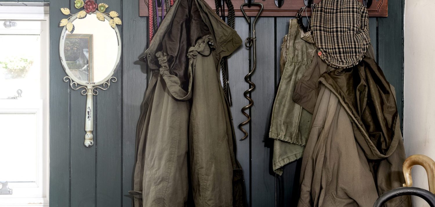 Coat rack close up on dark grey panelled wall with muted green coats hanging beside white antique mirror