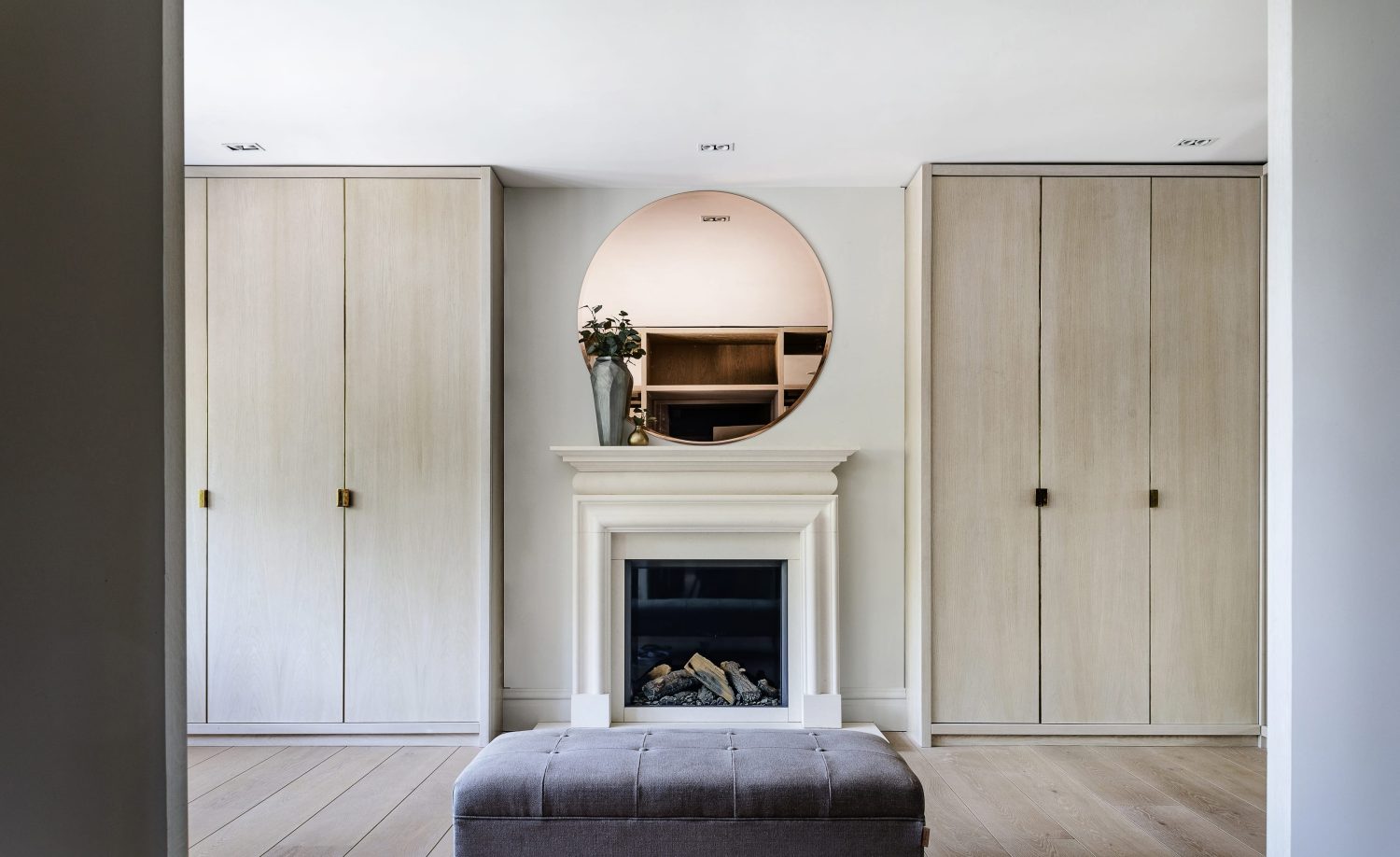 Dressing room area with wooden wardrobes on either side of a white fireplace with a large circular mirror on the wall above