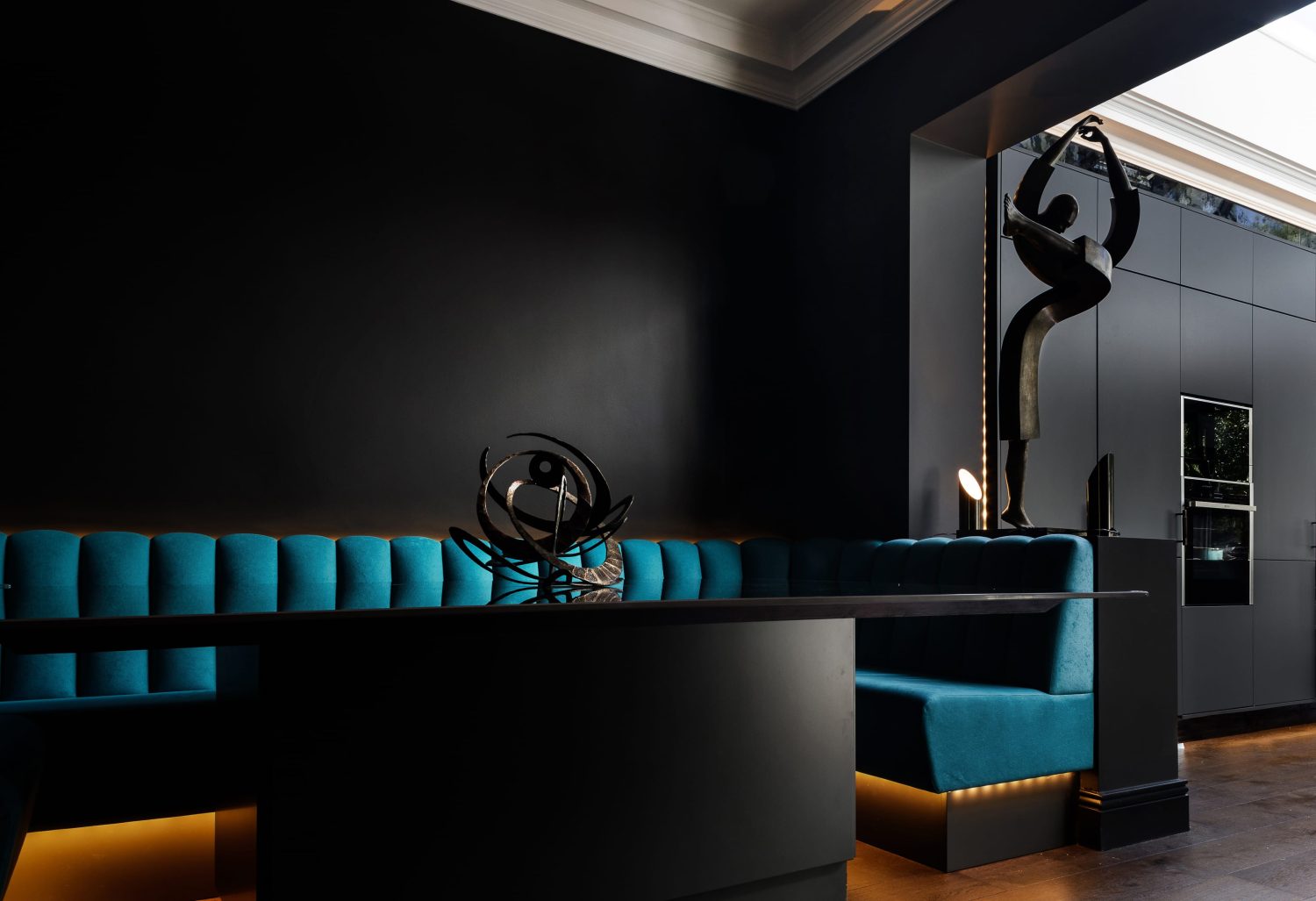 Black dining table with blue booth seating surrounding and a large abstract ornament on partition wall through to kitchen cabinets