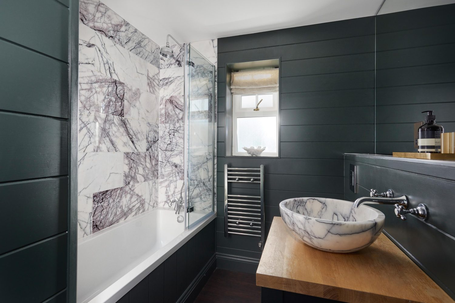 Bathroom with white and dark grey marble tiled walls around bath and dark panelled walls with chrome towel rack radiator and circular marble sink