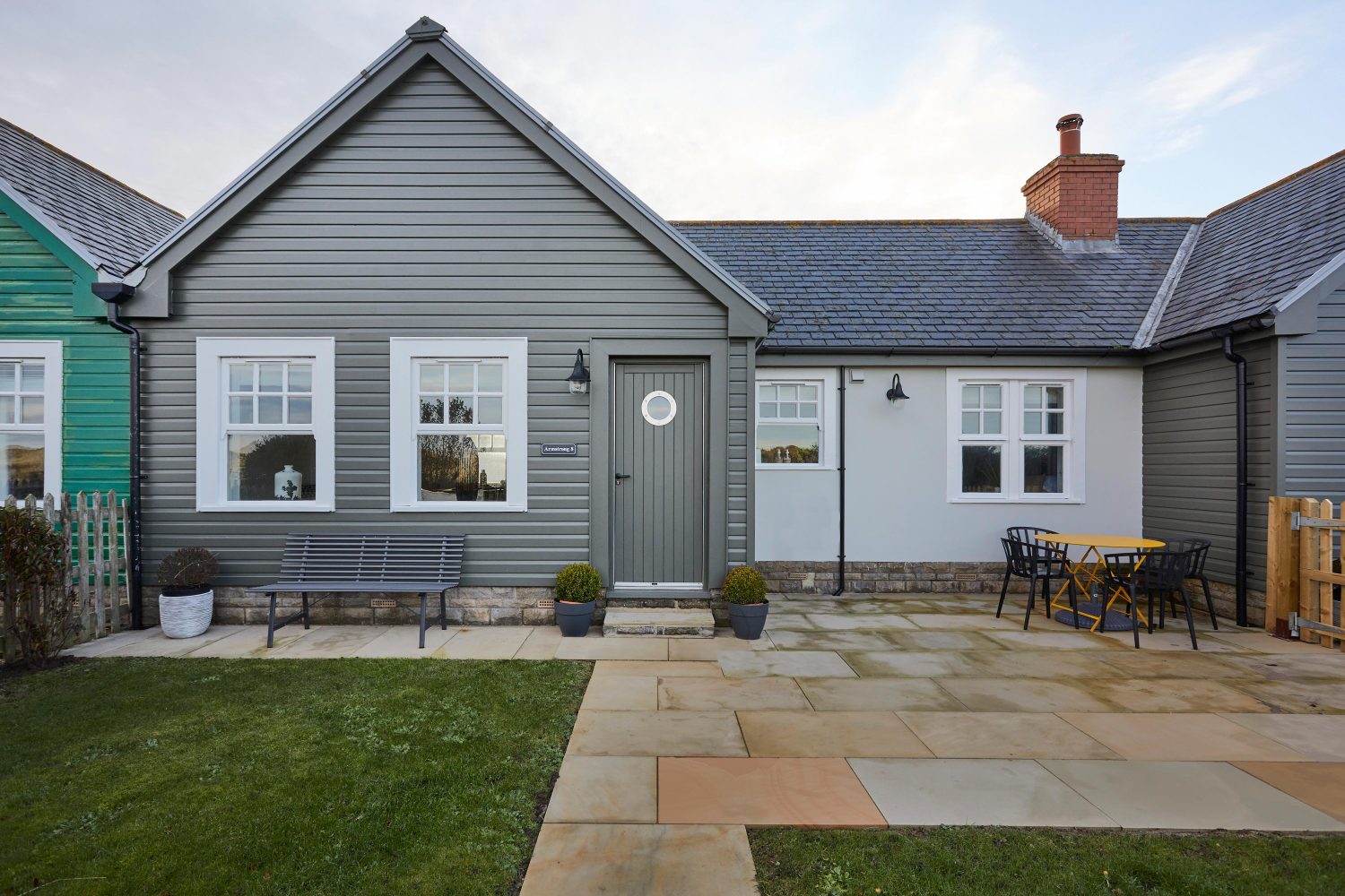 Exterior front of cottage with muted green panelled walls and door with stone paved walkway and outdoor seating
