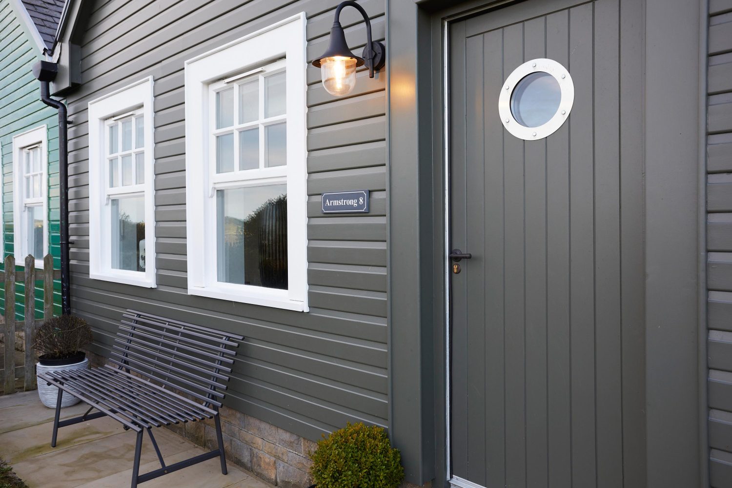 Exterior of cottage front door with muted green panelling and white framed windows above bench