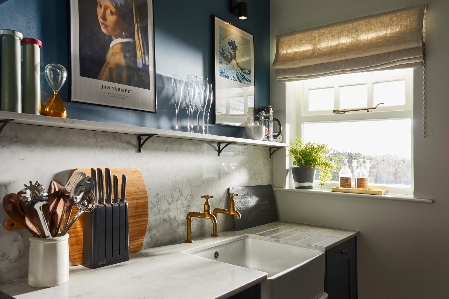 Kitchen with white sink and two gold taps and white marble countertop and backsplash with muted blue wall above and framed artwork