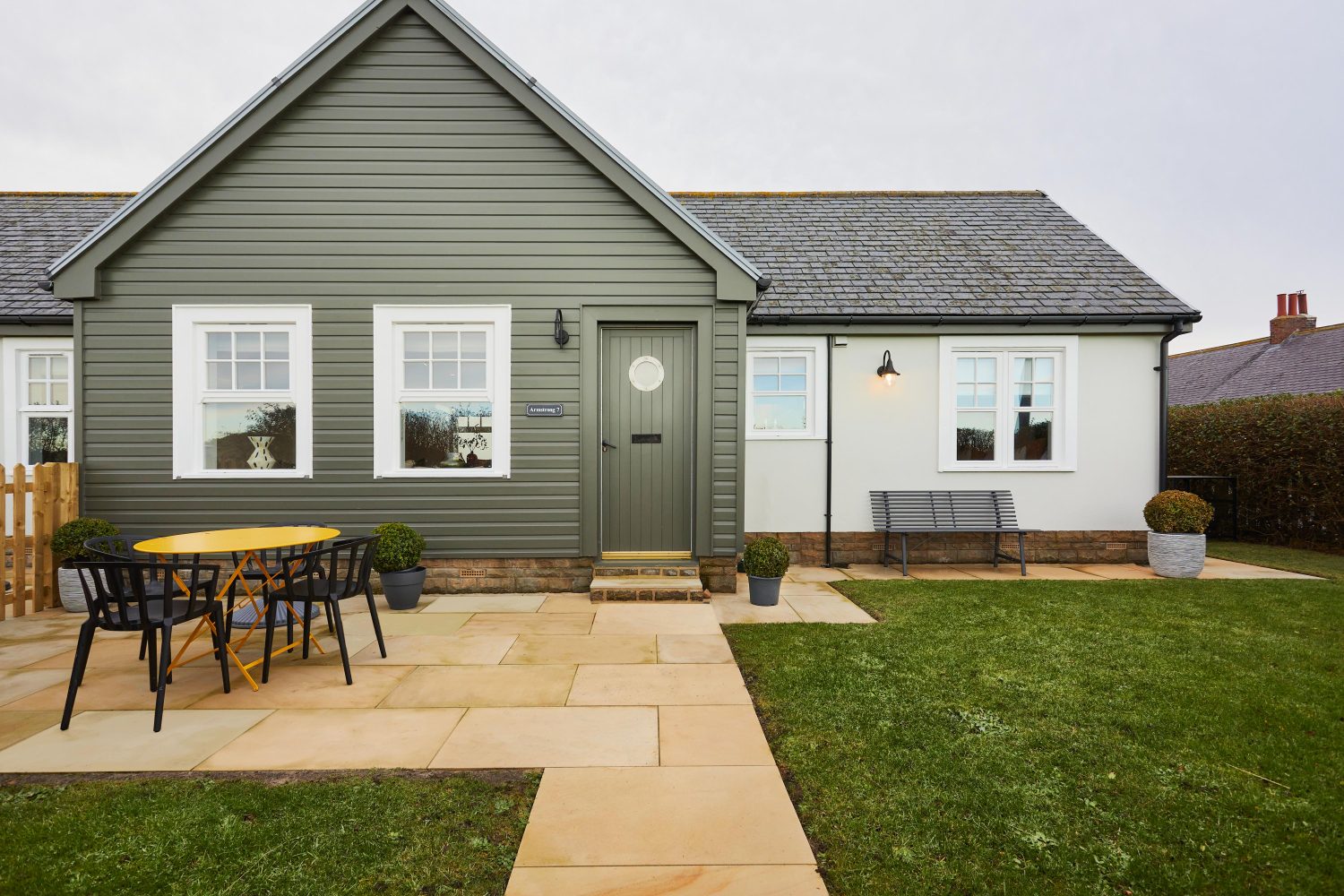 Exterior front of cottage with muted green panelled walls and door with stone walkway and grass in front