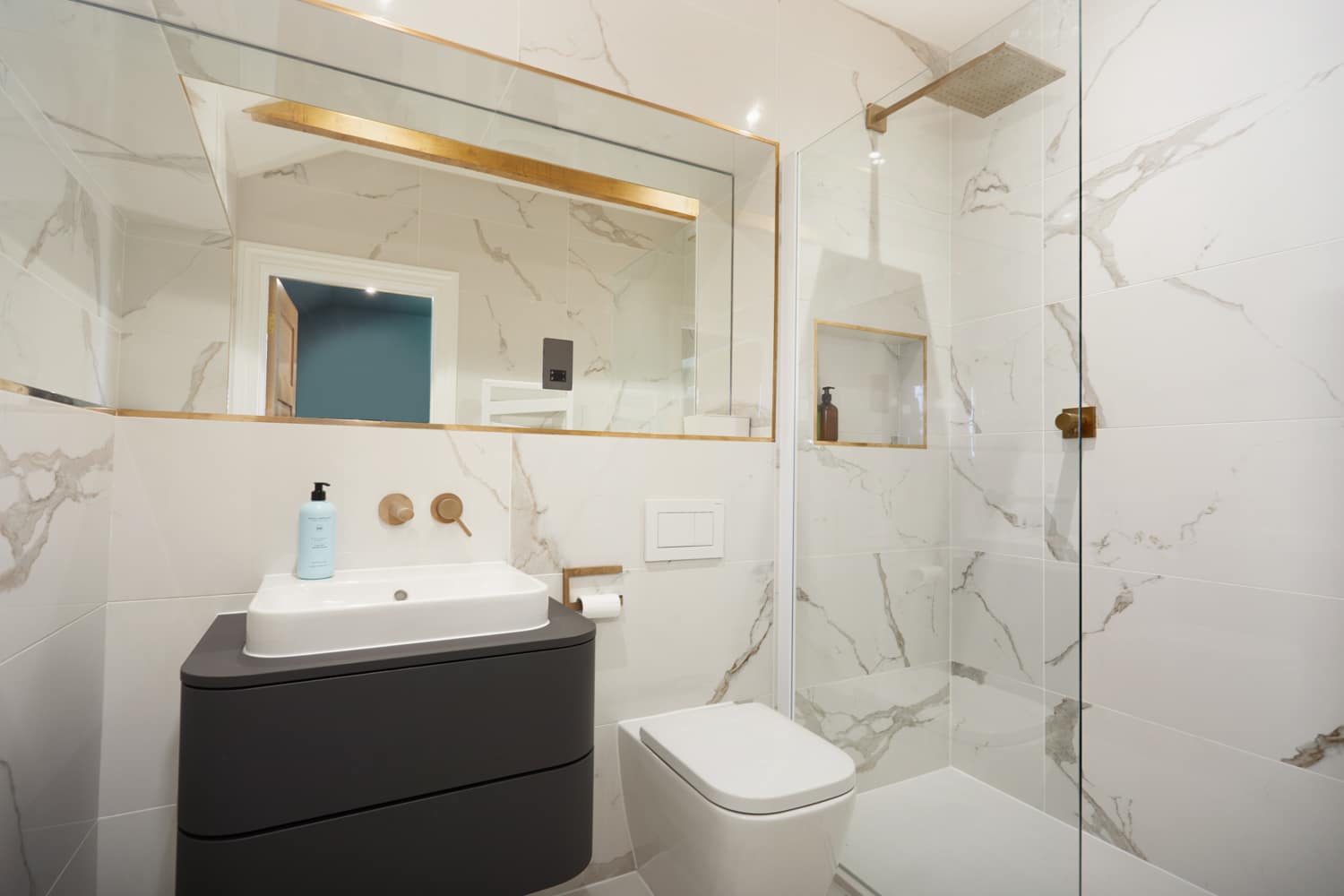 White marble bathroom with gold accents and large mirrr above dark grey sink unit