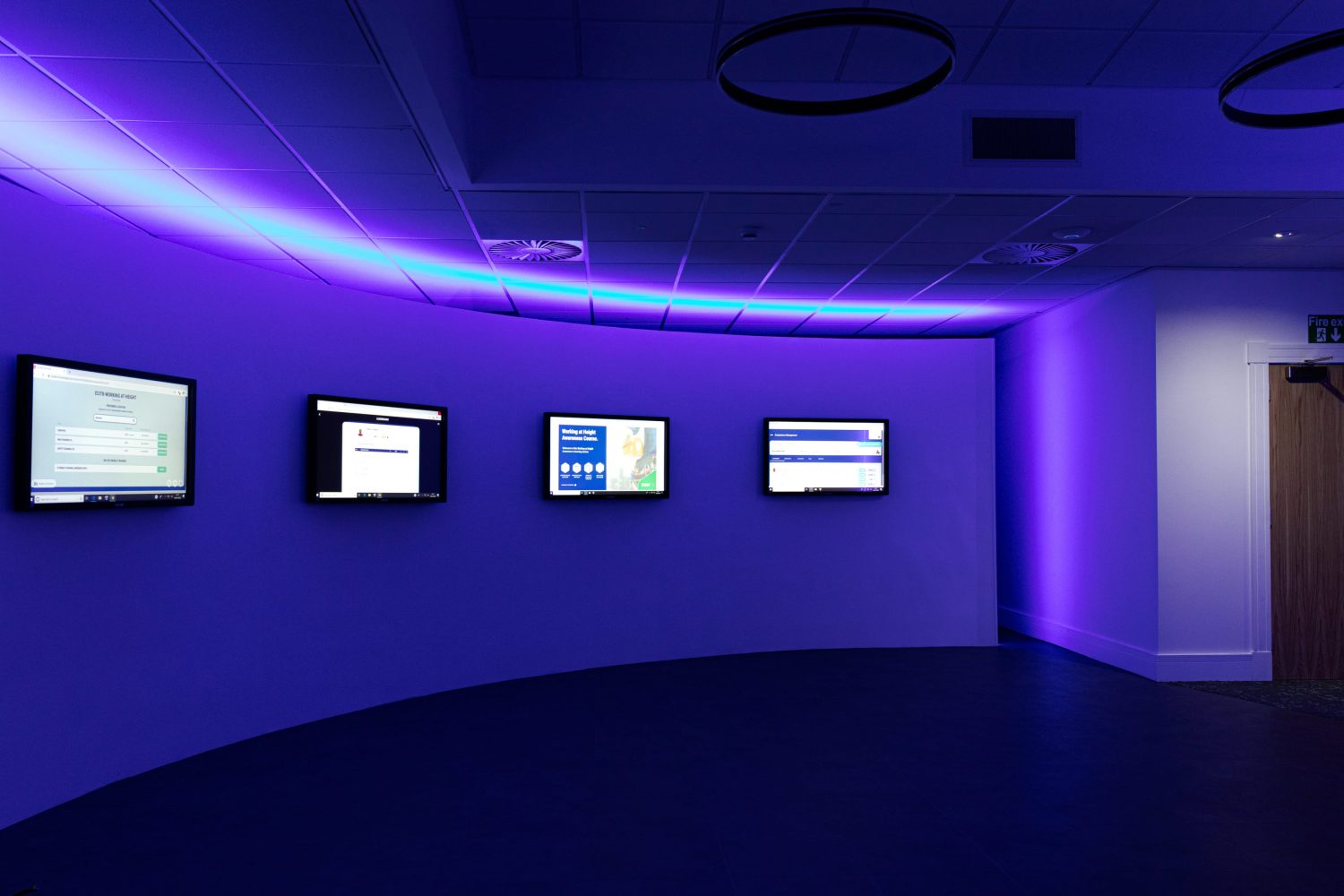 Four TVs mounted on curved wall with purple LEDs glowing above and circular pendants hanging from ceiling