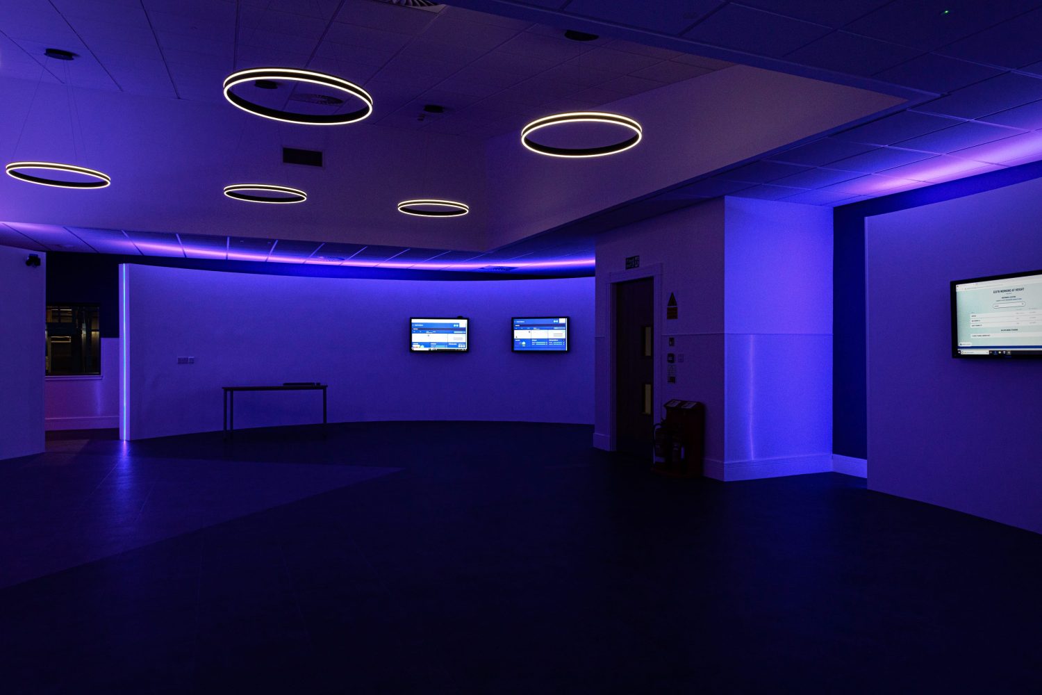 Room with TVs mounted to walls with purple glowing LEDs around and circular pendant lights hanging from ceiling