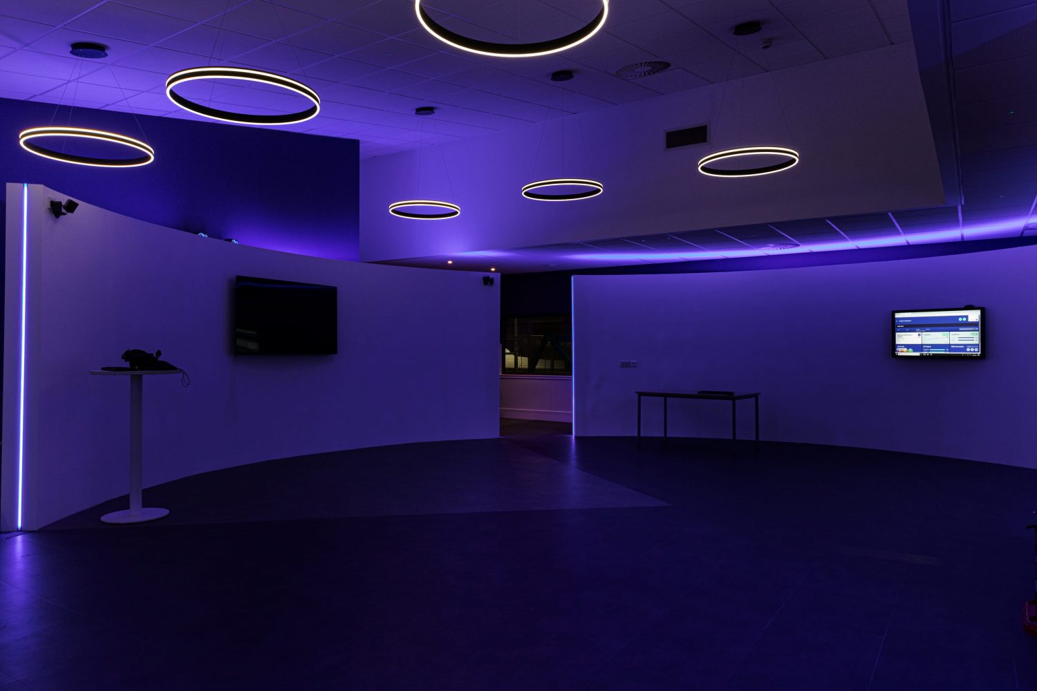 Room with two curved walls with TVs mounted and purple LEDs glowing around and circular pendant lights hanging from ceiling