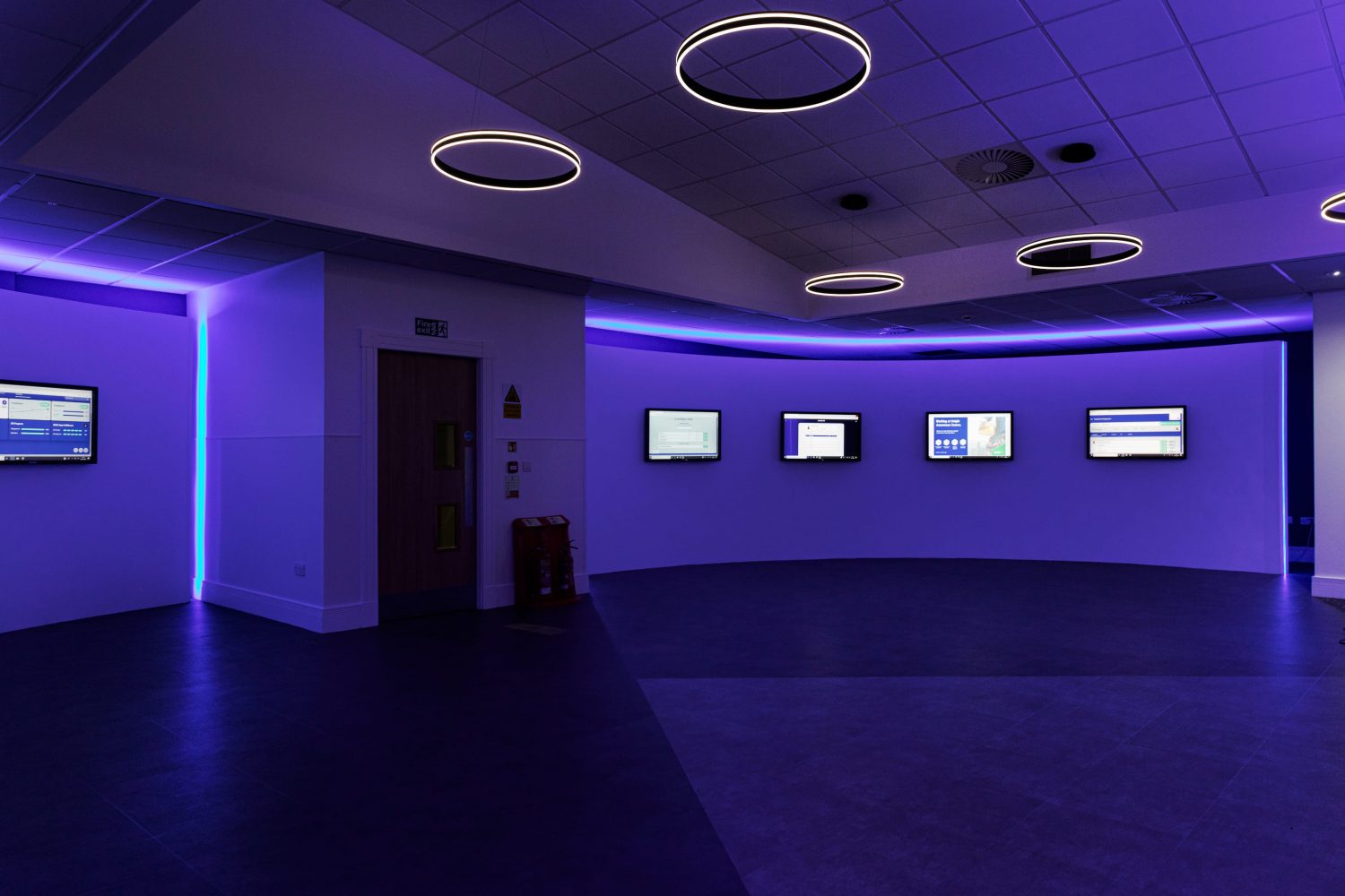 Room with curved wall with TVs mounted and purple LEDs glowing around and circular pendant lights hanging from ceiling