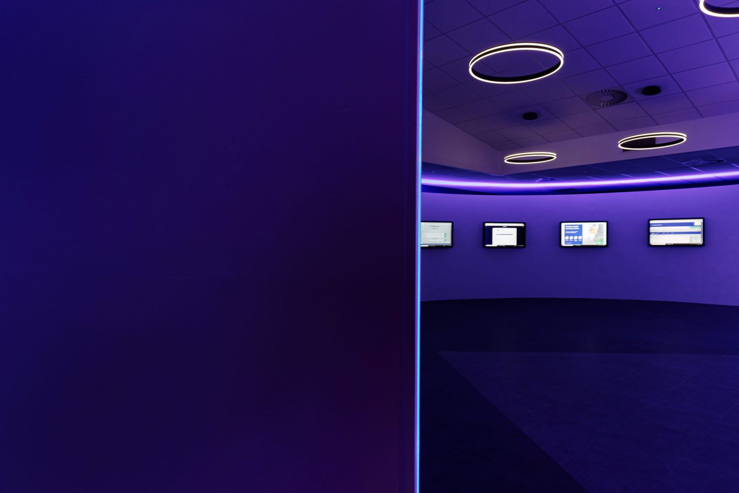 View from behind wall of TVs mounted to wall with purple LEDs glowing around and circular pendant lights hanging above