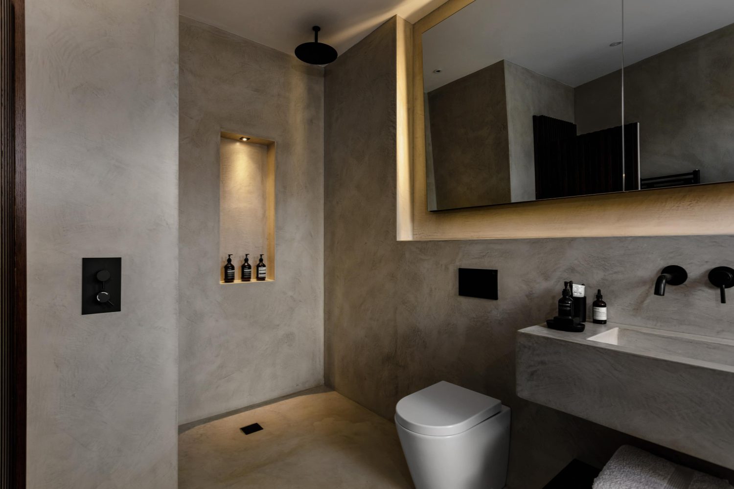 Bathroom with beige stone walls and large mirror on wall above built-in sink and tall shower niche with built-in spotlight to the side