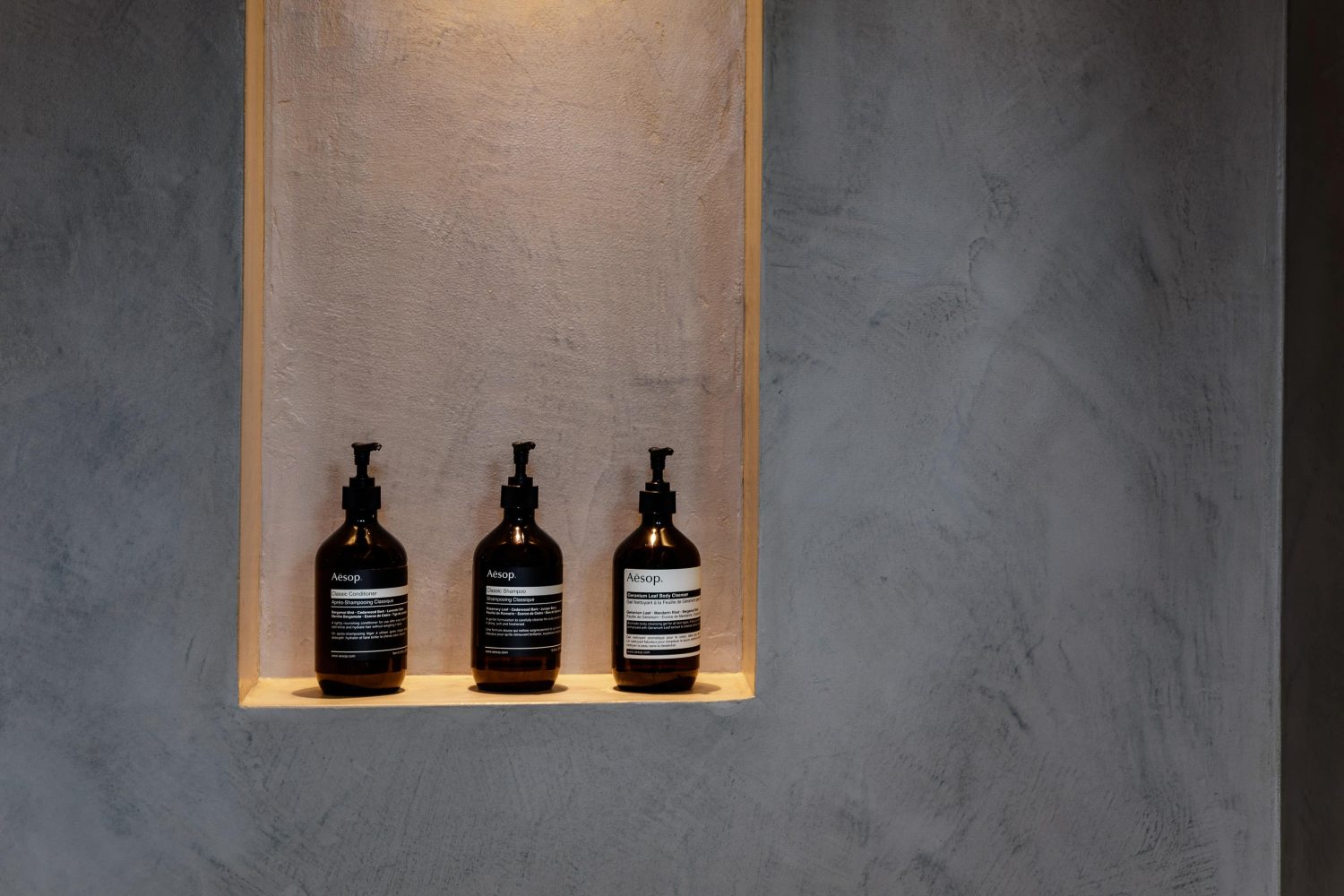 Close up on shower niche in a beige stone textured wall with a light from above and three bottles on the shelf