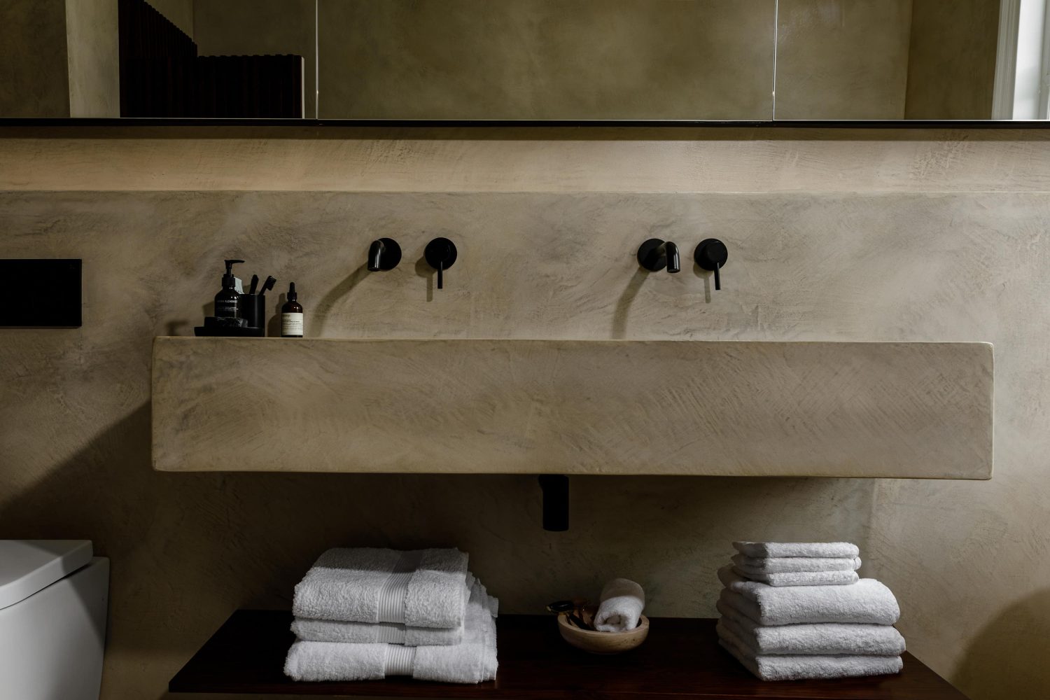 Stone bathroom sink built into wall with two set of black taps and faucets and white towels stacked on wooden shelf beneath