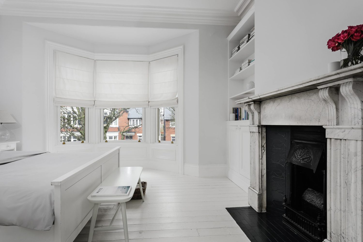 Bedroom with white double bed across from light grey marble and black fireplace with white walls surrounding and a large bay window on the far wall