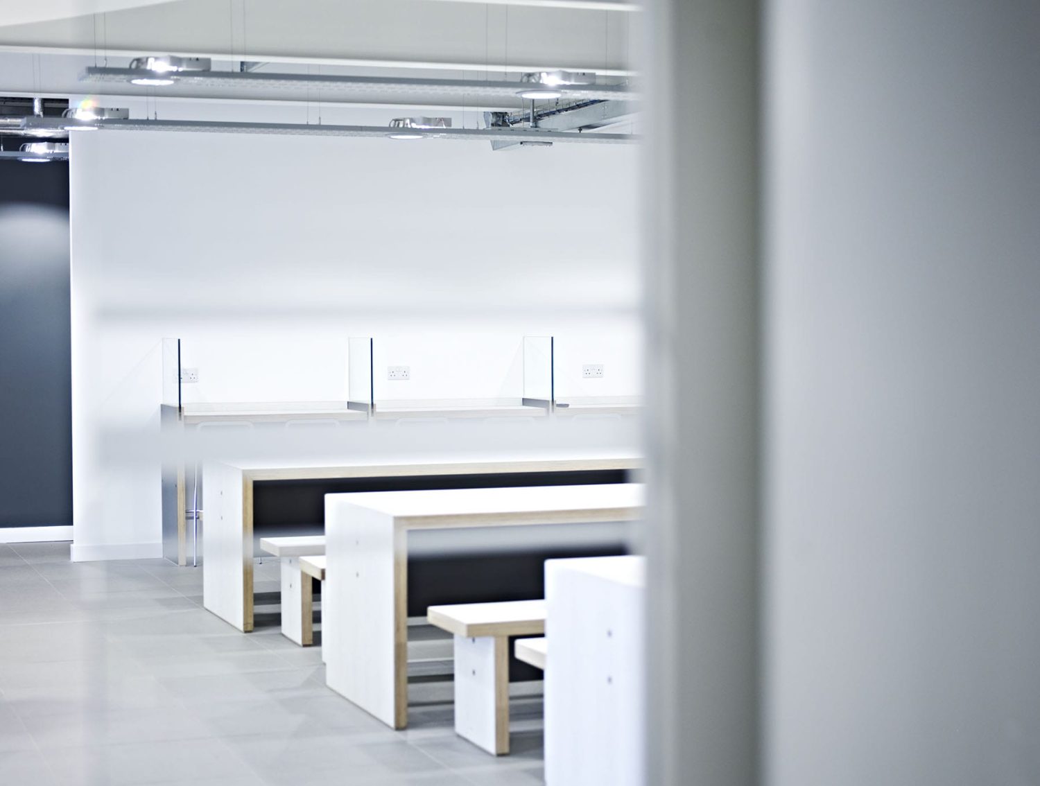 White desks in a row with benches beside and a white wall behind featuring a desk with glass partitions