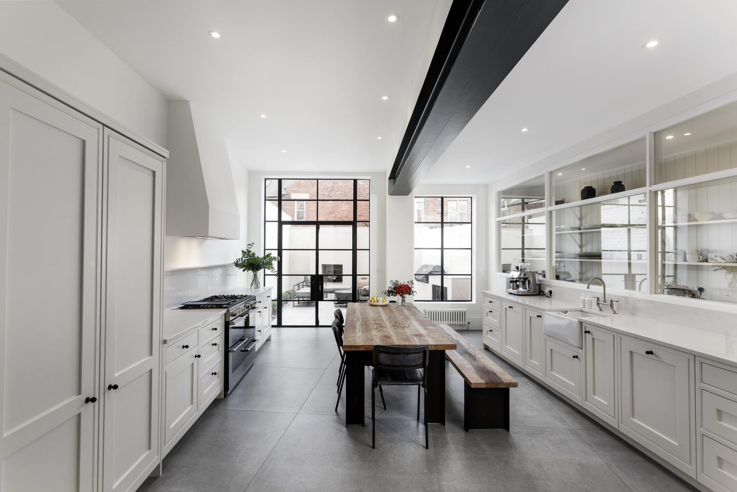 Kitchen with white walls and cabinets, Crittall-style double doors on the far wall and a rectangular wooden dining table with bench and chairs in the centre