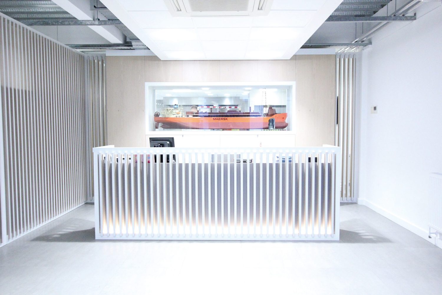 Reception desk with a large model ship encased in the wall behind. Partition walls of wooden slats around the front of the desk and behind it