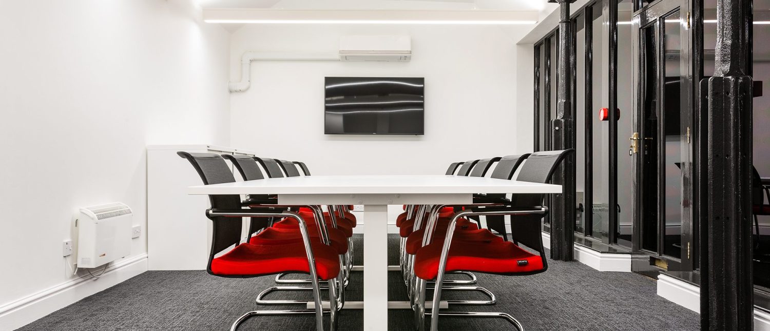 Meeting room with large white table in the centre and black and red seats around and a TV on the wall