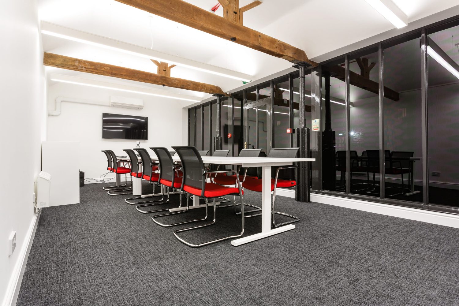 Meeting room with black framed windows across wall, large white table with black and red chairs and wooden beams across ceiling above
