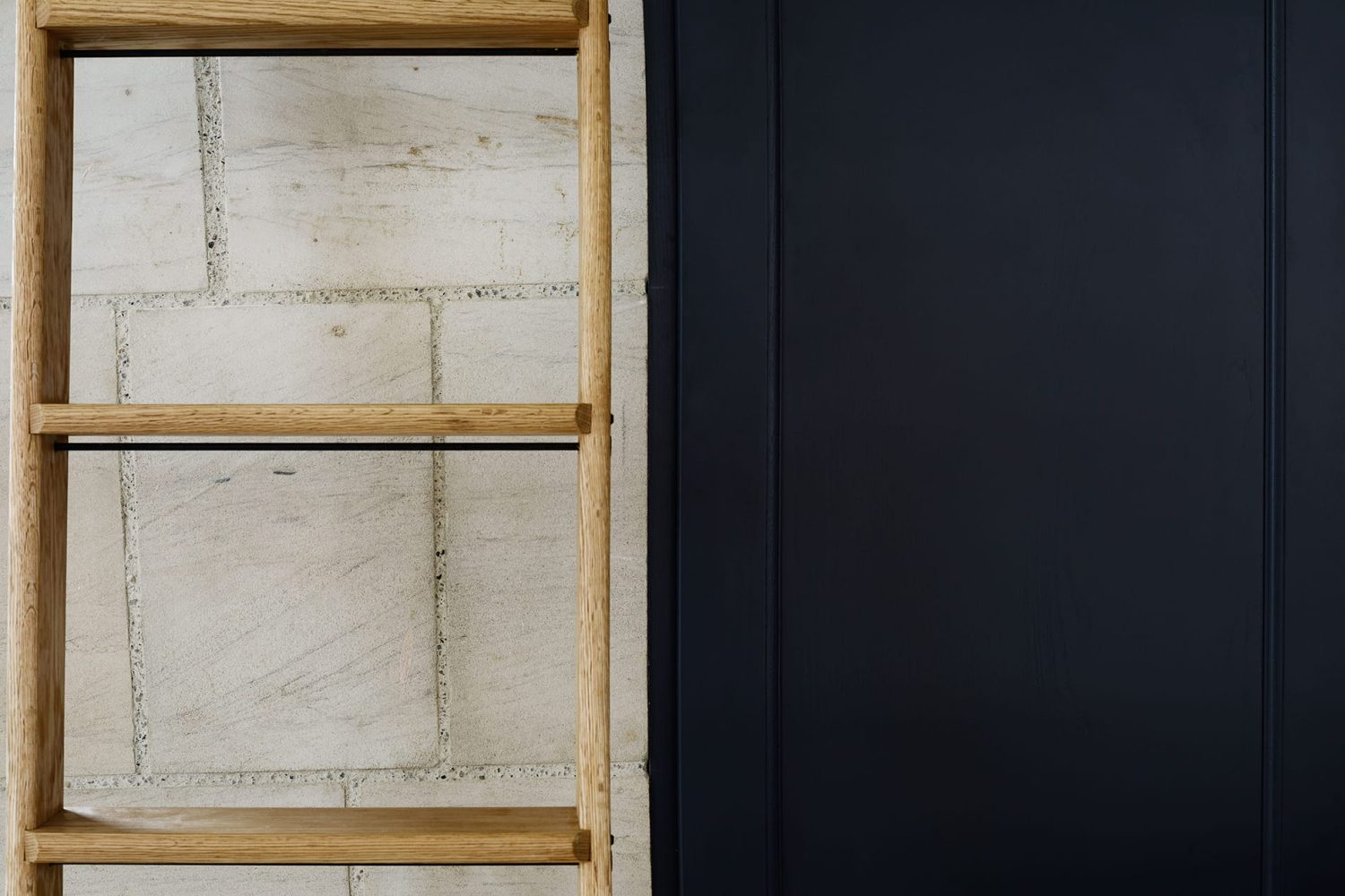 Close up wooden ladder beside wall of exposed stone and a black cabinet door