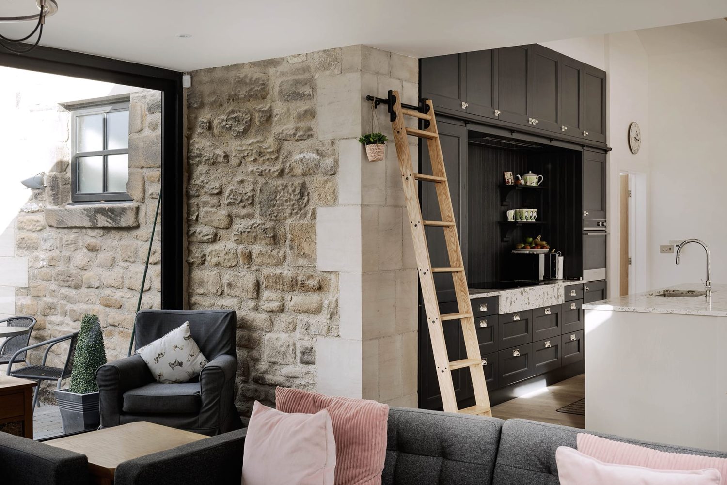 View of kitchen with black cabinets and wooden library ladder from living room with grey sofas