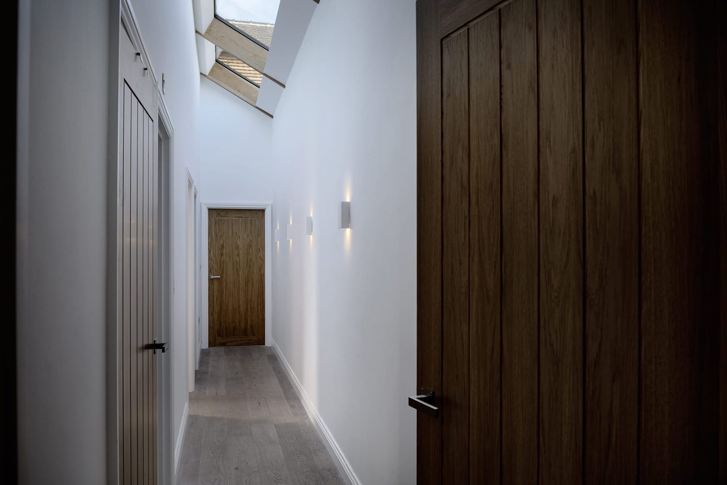 View through hallway with white walls and wooden doors with wall lamps and skylights above